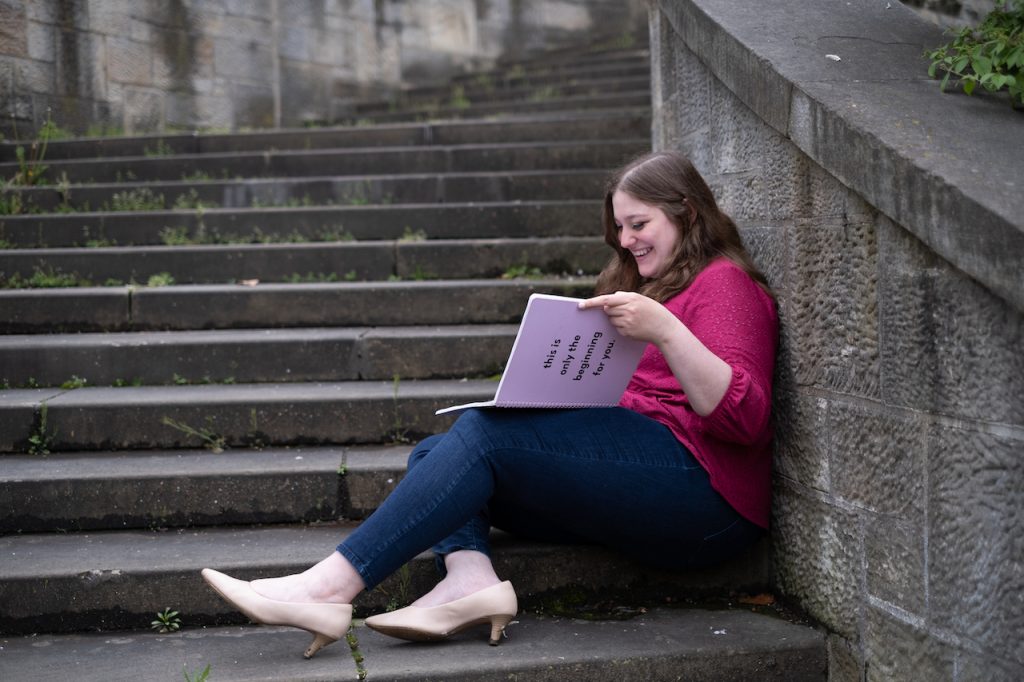 Rebecca Ferlotti in a pink top, jeans, and heels, sitting on steps with a pink journal open. The journal cover says, "This is only the beginning for you."