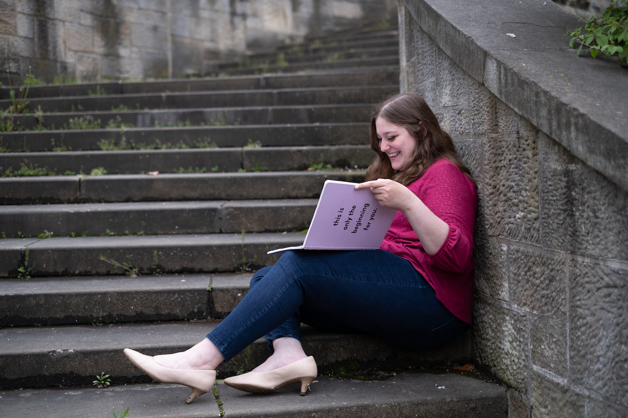 Rebecca Ferlotti in a pink top, jeans, and heels, sitting on steps with a pink journal open. The journal cover says, "This is only the beginning for you."